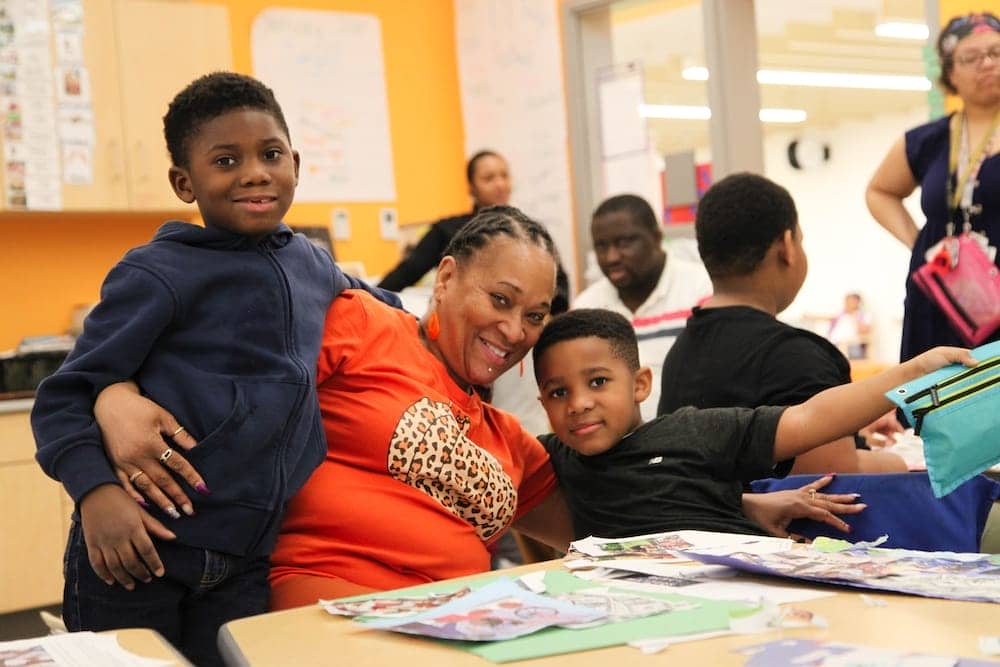 An adult in bright orange wearing a bright smile wraps their arms around two young students. They are at a table covered in paper materials in a bright gold and white classroom.