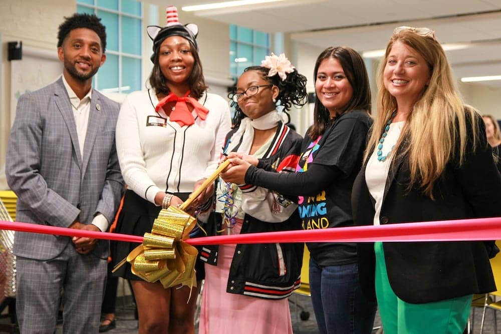 Baltimore Mayor Brandon Scott stands with Arts for Learning President and CEO Stacie Sanders Evans and three high-school students. All are smiling into the camera as the students brace a pair of giant golden scissors to cut a thick red ribbon and golden bow stretched out in front of them.